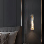 Lit Clérachic crystal pendant hanging beside a dark bed with grey headboard and cushions, in front of a charcoal wall with a bronze trim line.