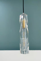 Faceted clear crystal shade of this Clérachic pendant, with its brass-toned cap and a filament bulb, against a teal wall.