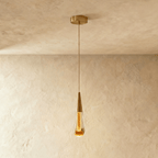 Clérachic single crystal drop pendant light with a brass cone cap, glowing warm amber against a plaster ceiling.