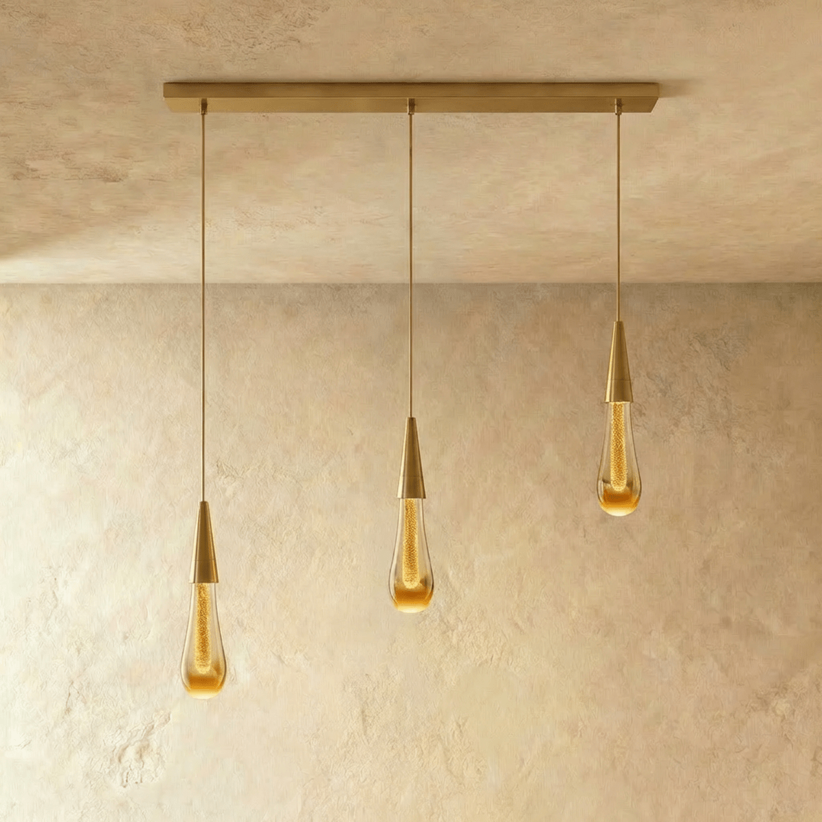 Clérachic crystal drop pendant light in a three-piece linear row, amber glass teardrops hung from a slim brass bar.
