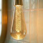 Close-up of a lit Clérachic crystal drop pendant's bubbled amber glass and brass cap against teal bathroom tile.