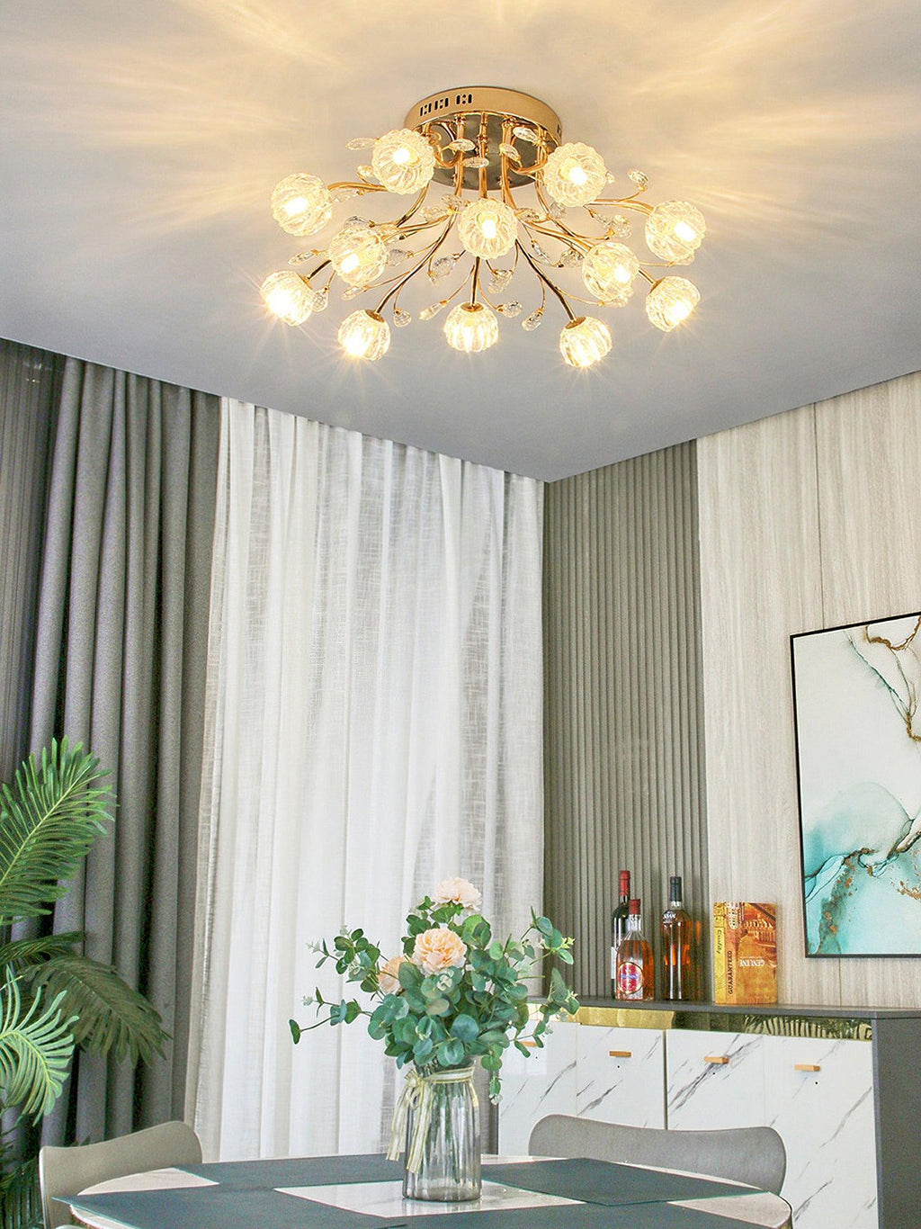 Gold ceiling light from Clérachic with glass flower shades lit above a dining table with a bouquet, in front of grey and white curtains.