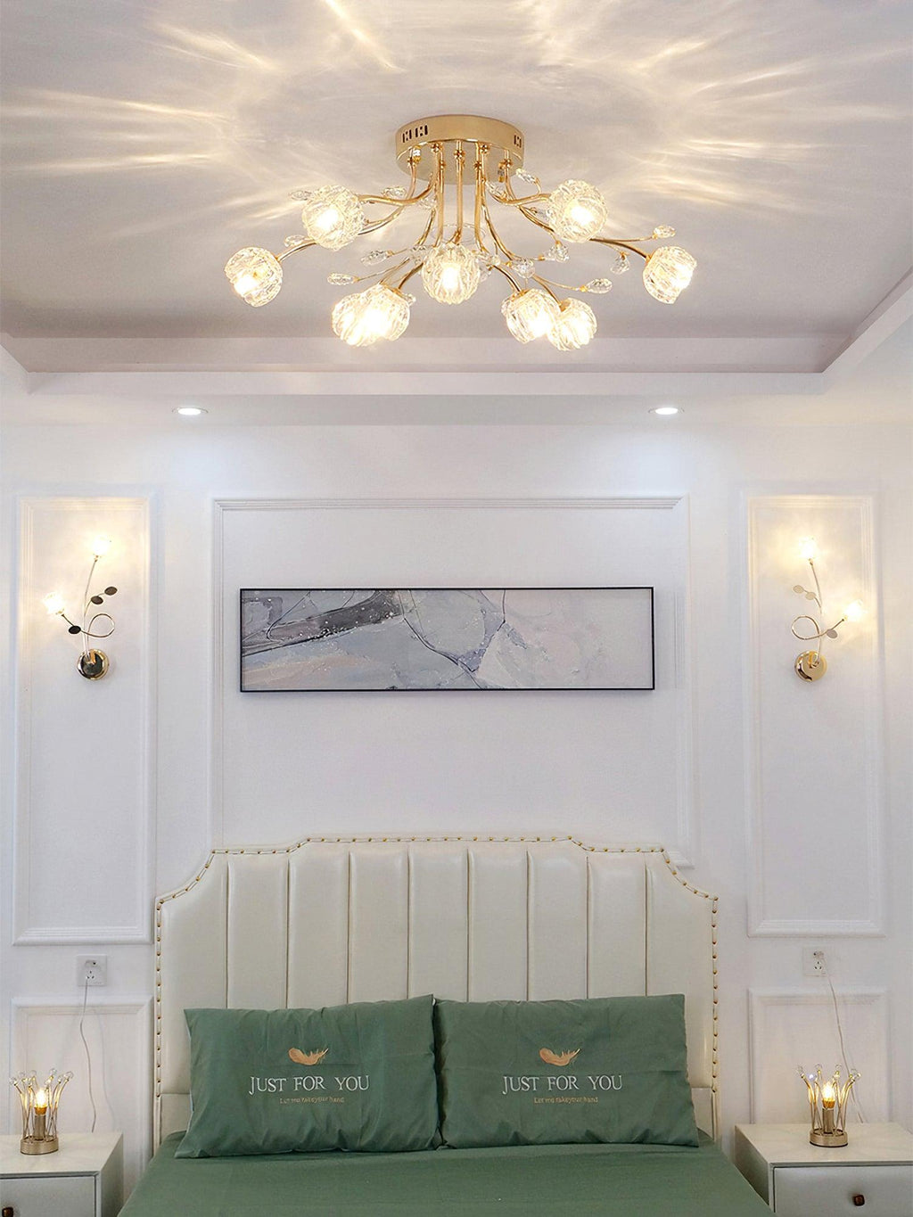 Clérachic gold ceiling light with crystal flower shades above a bed with a pale headboard, green cushions and two wall sconces.