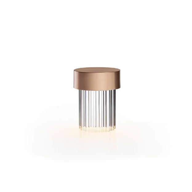 A Clérachic lamp with a rose gold cap and fluted clear glass on white, with a warm glow spreading from its base.