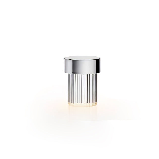 A Clérachic lamp with a polished chrome cap and fluted glass on white, glowing softly at the base.