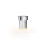 A Clérachic lamp with a polished chrome cap and fluted glass on white, glowing softly at the base.