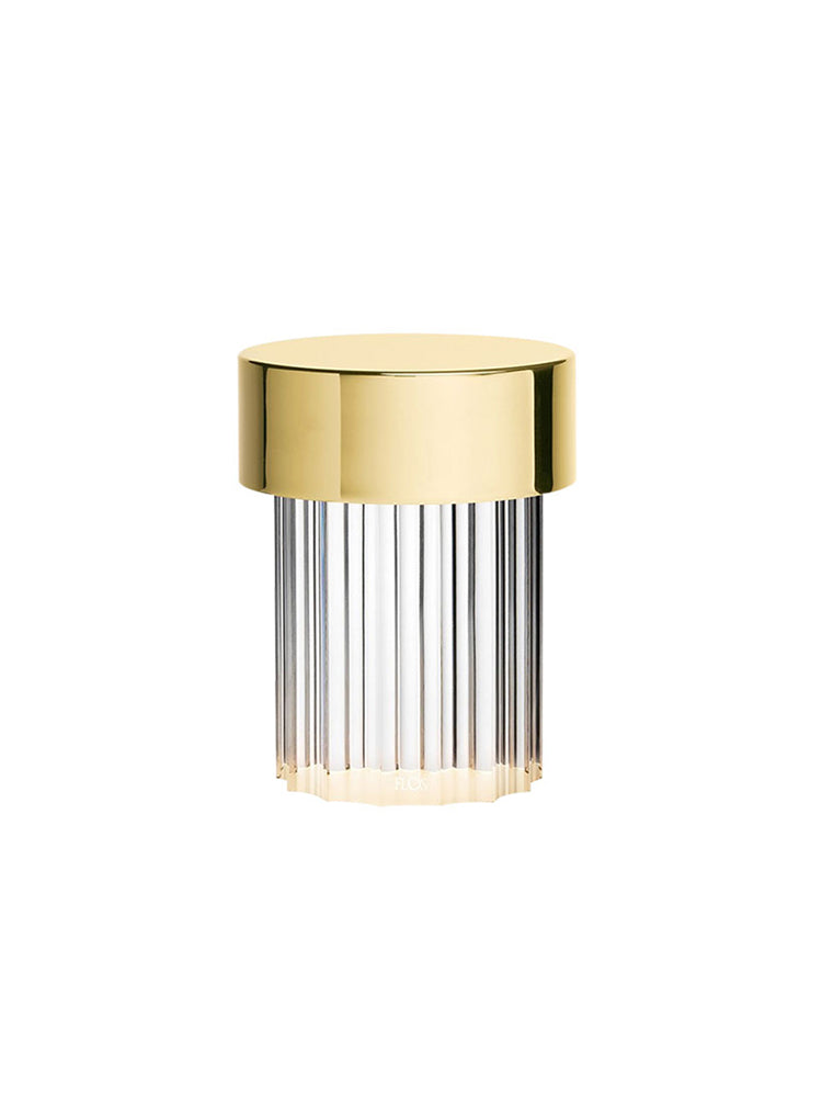 A fluted-glass Clérachic lamp with a polished gold cap on white, with the base lit a warm white.