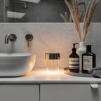 A Clérachic lamp with a chrome cap and fluted glass glowing on a marble bathroom counter beside a basin and dried grasses.