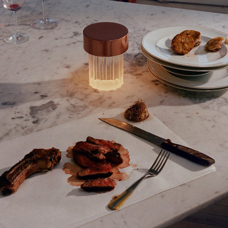 A fluted Clérachic lamp with a copper-toned cap lit on a marble counter, next to a plate of meat and a knife and fork.