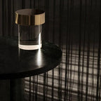 A gold-capped Clérachic lamp with a clear glass base on a dark round table, with fine dark strings hanging behind it.