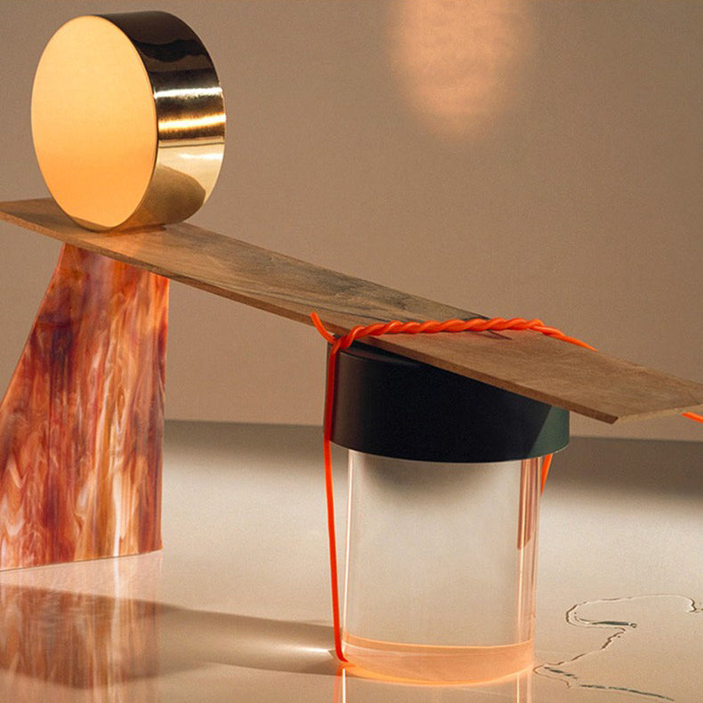 Close view of a Clérachic lamp under a wooden plank, with a gold disc balanced on top and a strand of orange cord.