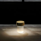 A fluted Clérachic lamp with a gold cap glowing in a dark room, its light spreading in a pool across a pale surface.