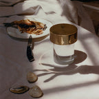 A smooth-glass Clérachic lamp with a gold cap on a white cloth beside a plate of fish, a knife and a few shells.