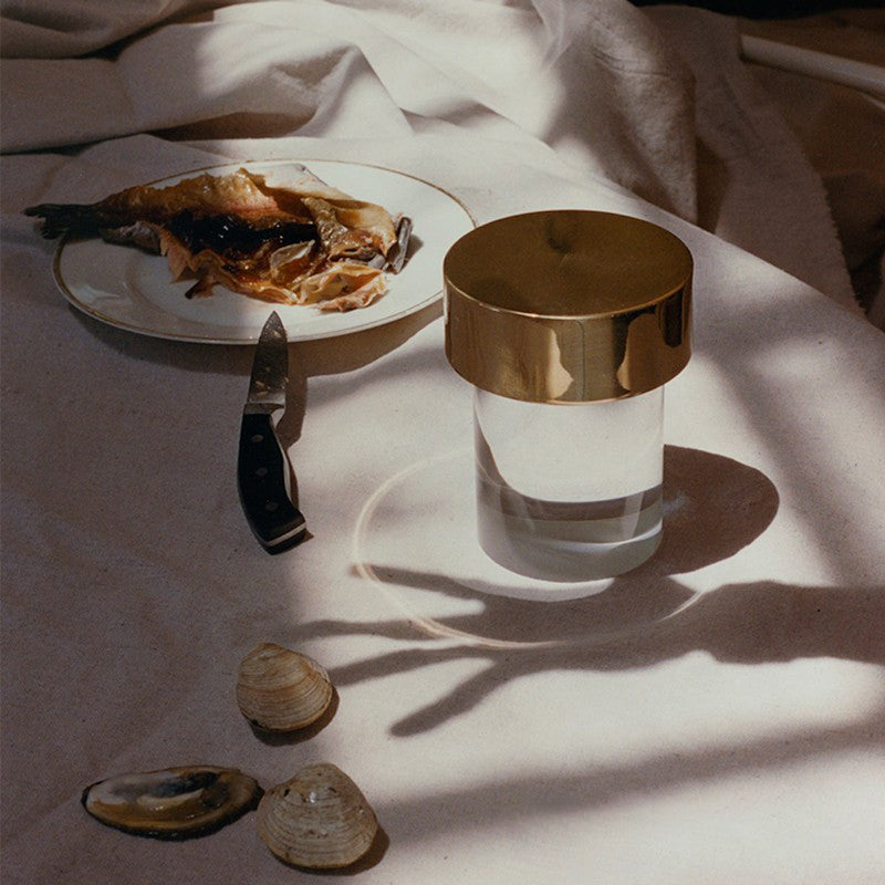 A smooth-glass Clérachic lamp with a gold cap on a white cloth beside a plate of fish, a knife and a few shells.