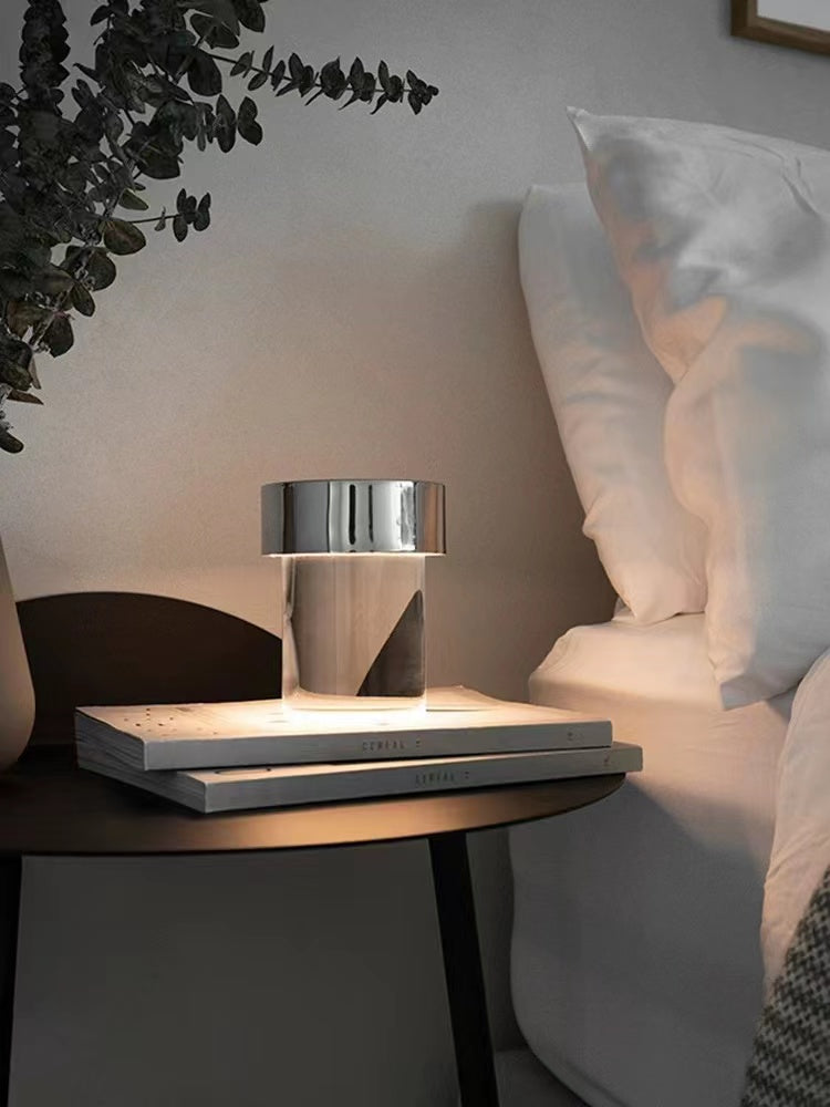 A Clérachic lamp with a chrome cap on stacked books on a dark bedside table, with a white pillow and leaves behind it.