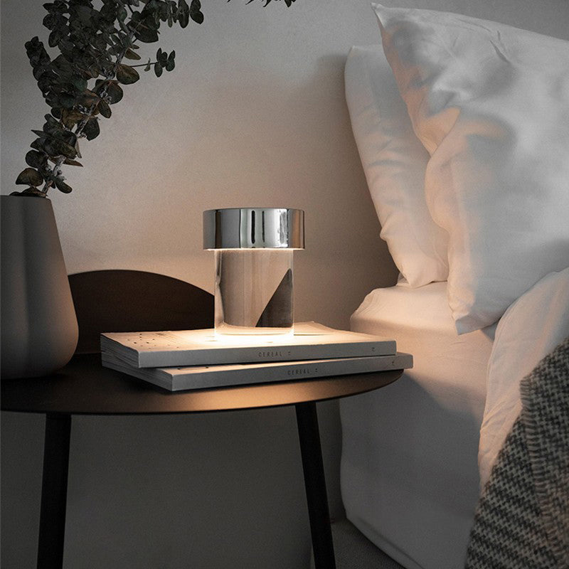 A lit Clérachic lamp with a chrome cap on two books on a round dark bedside table, beside a grey vase and white bedding.