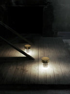 Tall crop of two Clérachic lamps glowing on a pale wooden floor in a dark room, casting light across the boards.