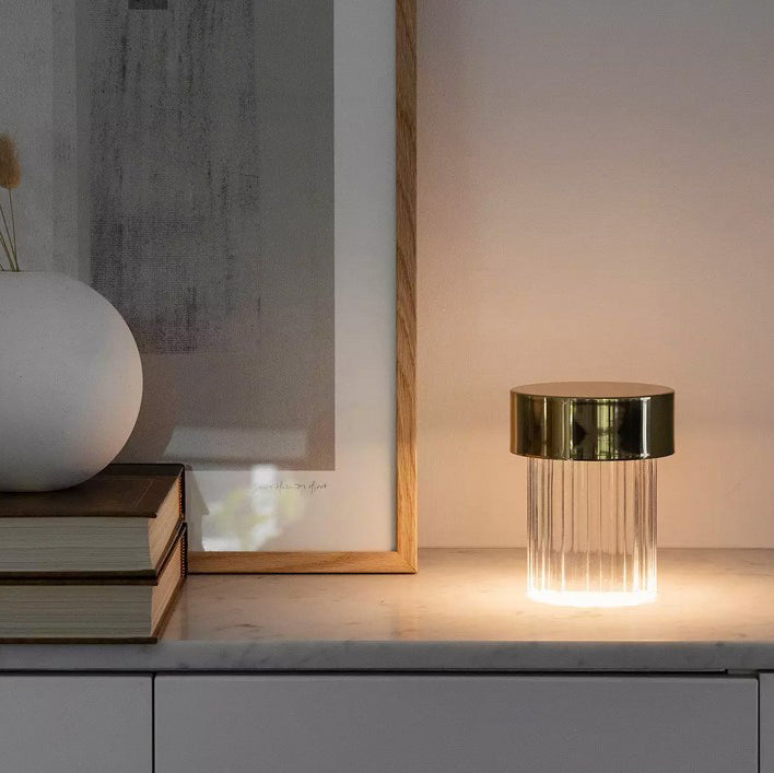 A fluted Clérachic lamp with a gold cap glowing on a marble sideboard, beside books, a round white vase and a framed print.