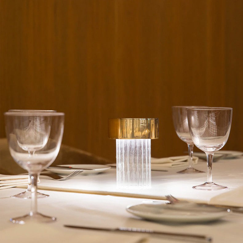 A fluted Clérachic lamp with a gold cap lit on a restaurant table among wine glasses, cutlery and a plate, with a wood-panelled wall behind.