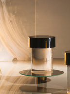 A Clérachic lamp with a black cap and clear glass sitting on a round glass tray in a warm, brushstroke-lit set.