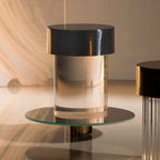 Close view of a Clérachic lamp with a black cap and clear glass body on a round glass tray, with warm light behind.
