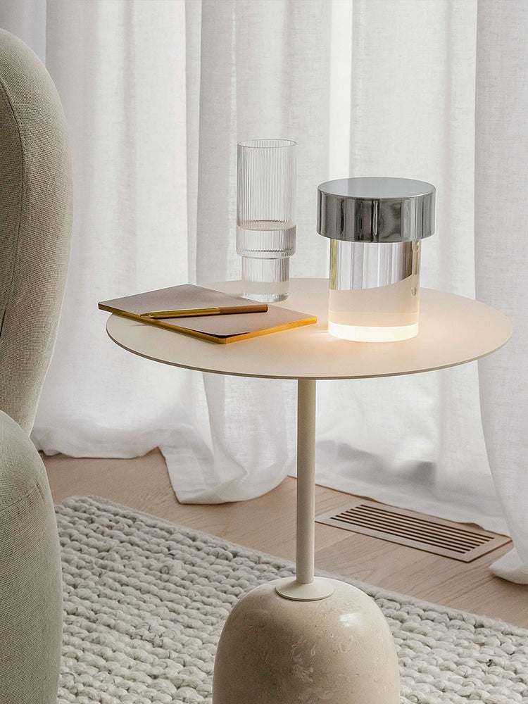 A Clérachic lamp with a chrome cap glowing on a small cream side table by white curtains, with a notebook and a ribbed glass beside it.