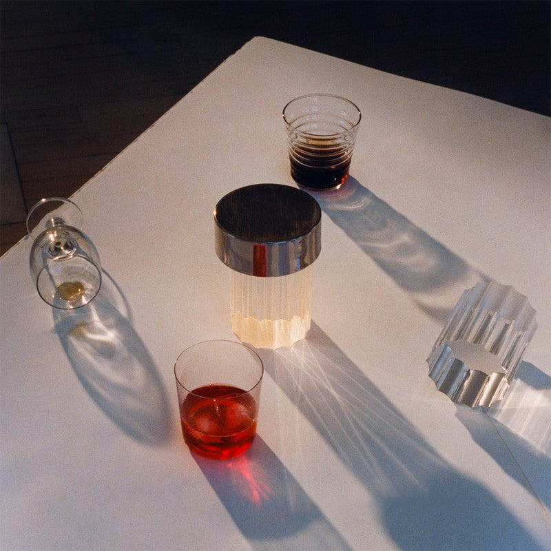 Looking down on a fluted Clérachic lamp with a chrome cap on a white table, with glasses of red and brown drinks and long shadows.