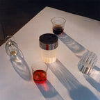 Looking down on a fluted Clérachic lamp with a chrome cap on a white table, with glasses of red and brown drinks and long shadows.