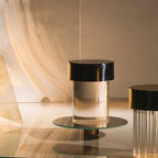 A black-capped Clérachic lamp on a round glass tray, with a second fluted lamp at the right edge and warm, streaked light behind.
