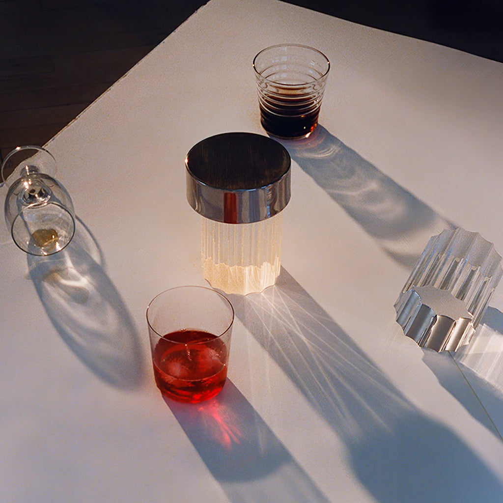 Overhead view of a fluted Clérachic lamp with a chrome cap glowing on a white table, beside two glasses of drink and sharp shadows.