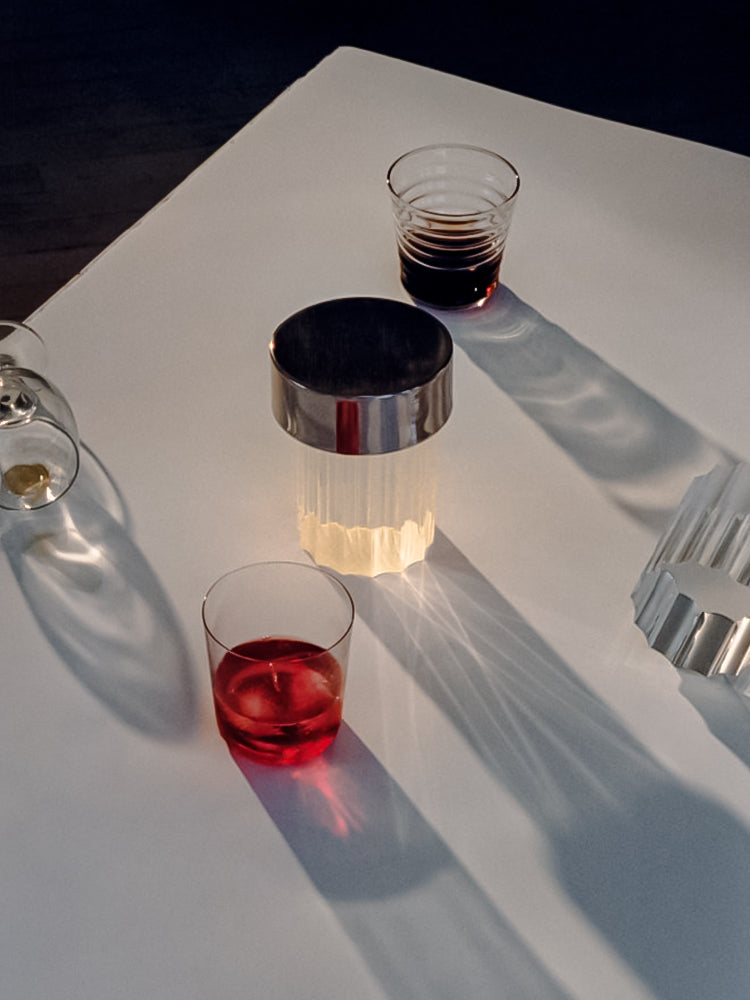 A fluted Clérachic lamp with a chrome cap on a white table, with a glass of red drink and a glass of brown drink, and sharp shadows.