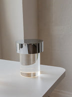 A Clérachic lamp with a chrome cap and smooth clear glass on a white table by a plain pale wall.