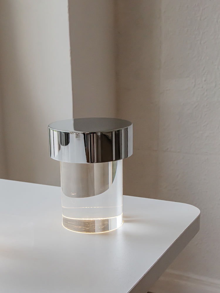 A Clérachic lamp with a chrome cap and smooth clear glass on a white table by a plain pale wall.