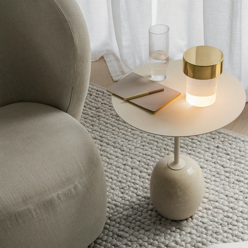 Looking down on a Clérachic lamp with a gold cap lit on a round cream side table, beside a notebook, a glass and a soft boucle chair.