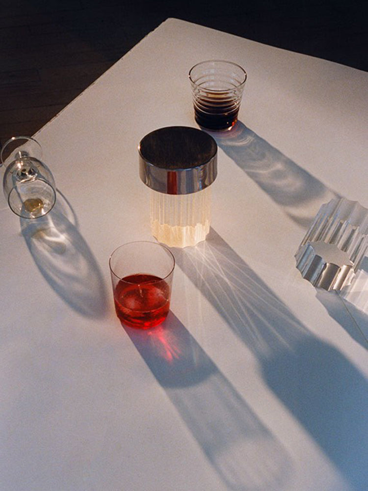 A fluted Clérachic lamp with a chrome cap lit on a white table, with two glasses of drink and long shadows across the surface.
