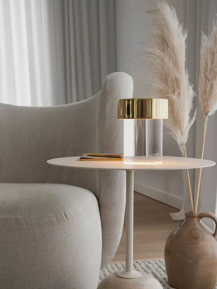 A Clérachic lamp with a gold cap and smooth glass on a slim white side table beside a cream armchair and pampas grass.