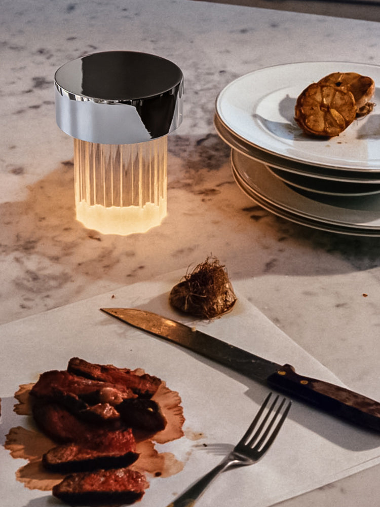 A Clérachic lamp with a chrome cap and fluted glass glowing on a marble counter, beside plates, a knife and a fork.