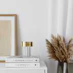 A fluted Clérachic lamp with a gold cap on two books on a white sideboard, beside a framed print and dried pampas grass.