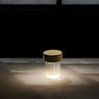 A fluted Clérachic lamp with a gold cap glowing in the dark, lighting a wide pool on the surface it stands on.