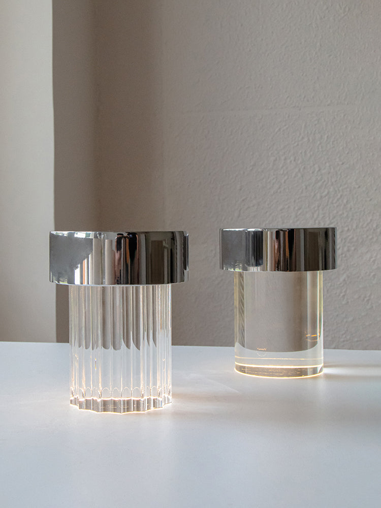 Two Clérachic lamps with chrome caps lit side by side on a white table, one with fluted glass and one with smooth glass.