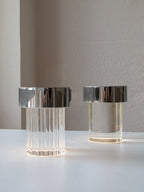 Two Clérachic lamps with chrome caps lit side by side on a white table, one with fluted glass and one with smooth glass.