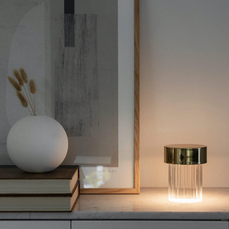 A fluted Clérachic lamp with a gold cap glowing on a marble sideboard, with books, a round white vase and dried stems beside it.