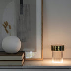 A fluted Clérachic lamp with a gold cap glowing on a marble sideboard, with books, a round white vase and dried stems beside it.