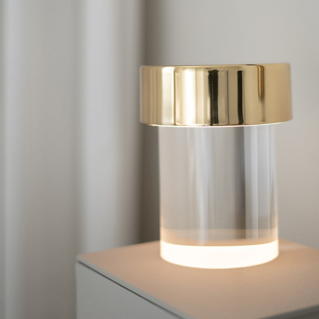 Close view of a Clérachic lamp with a gold cap, its smooth glass base glowing warm white on a pale plinth.
