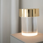 Close view of a Clérachic lamp with a gold cap, its smooth glass base glowing warm white on a pale plinth.
