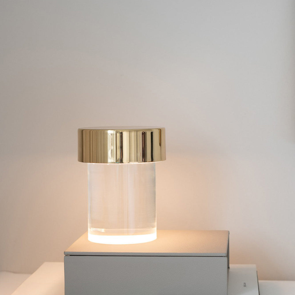 A Clérachic lamp with a gold cap and smooth glass lit on a white box against a plain grey wall.