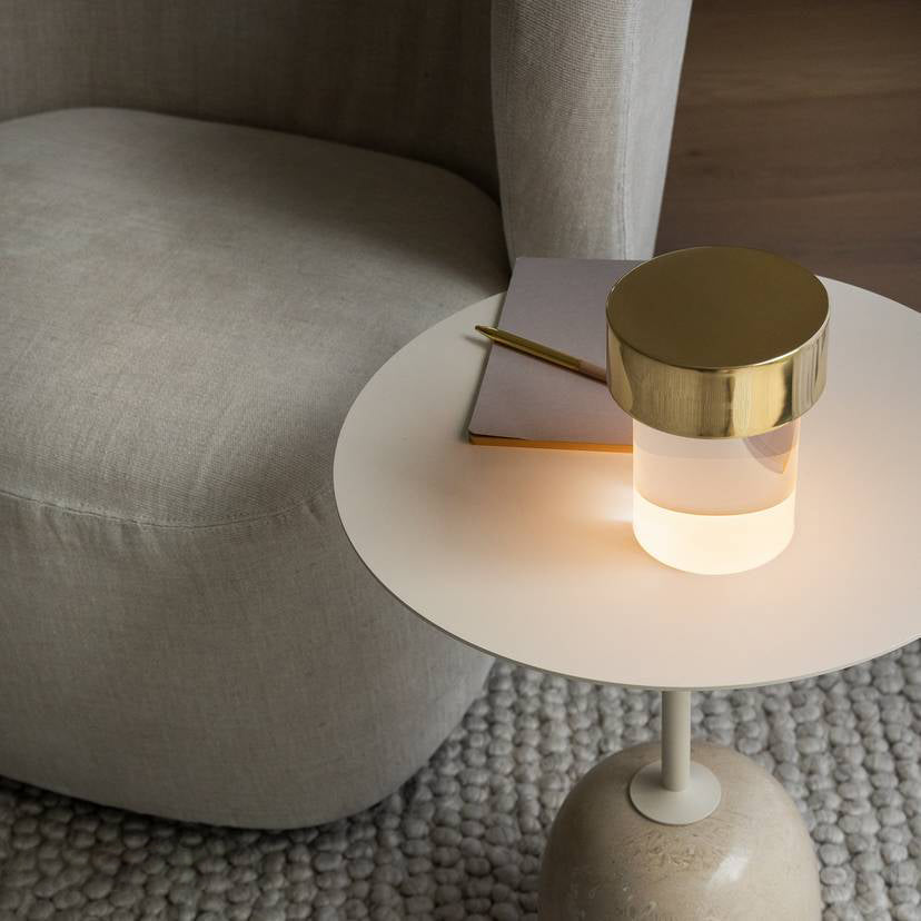 Looking down on a Clérachic lamp with a gold cap glowing on a round cream side table, beside a notebook and a pale sofa.