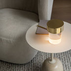 Looking down on a Clérachic lamp with a gold cap glowing on a round cream side table, beside a notebook and a pale sofa.