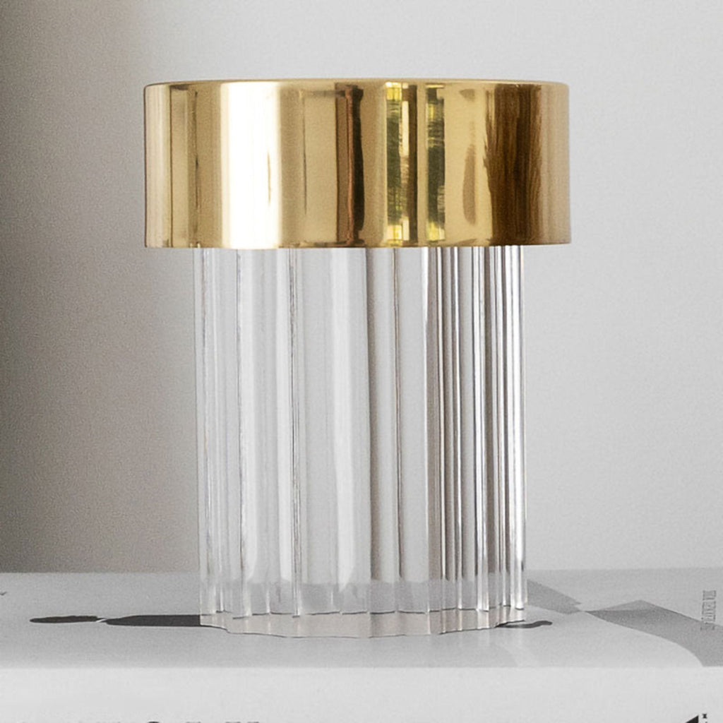 Close view of a fluted Clérachic lamp with a wide polished gold cap, standing on a book on a pale shelf.