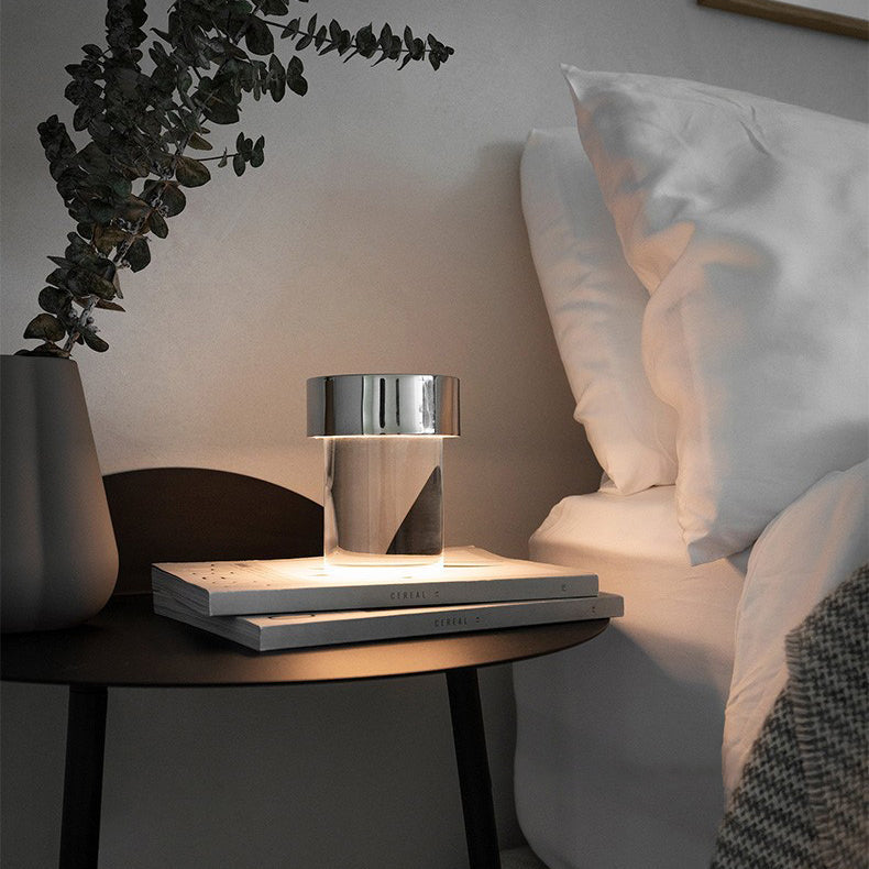 A Clérachic lamp with a chrome cap lit on two books on a dark bedside table, with white pillows and leaves behind it.